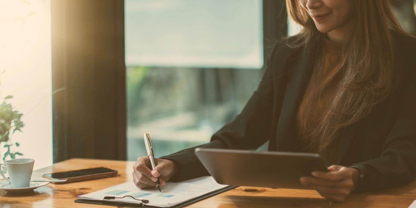 Businesswoman working on an enterprise grc program, holding a tablet and writing on a clipboard in a sunlit office.