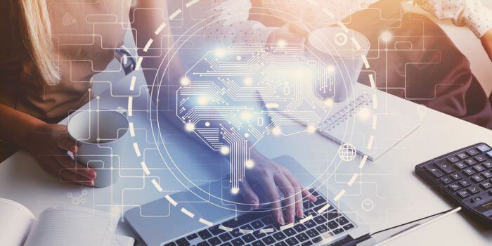 Woman sitting at desk with one hand typing on laptop and calculator in frame; a digital icon of a brain in a circle is over the laptop representing how regtech transforms regulatory compliance management