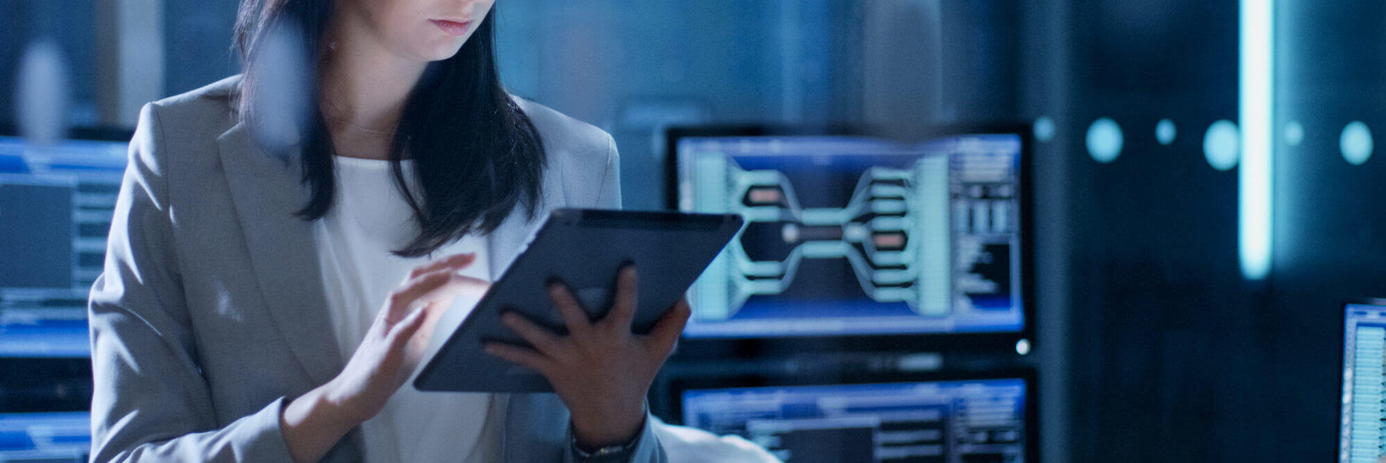 Young female government employee wearing glasses uses tablet in system control center. In the background her coworkers are at their workspaces with many displays showing valuable data.