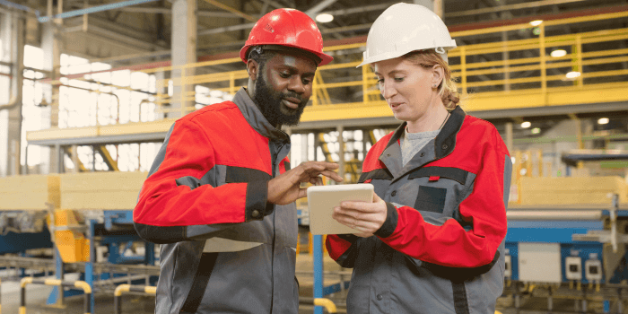 Woman in hardhat wearing ppe standing beside a man in a hardhat and ppe holding tablet representing manufacturing security