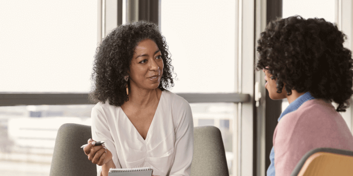 Two professional women in a conversation, seated across from each other in a bright office setting. One woman is holding a pen and notepad, attentively listening and taking notes, while the other speaks. The background features large windows with a view of a cityscape, suggesting a formal meeting or interview context representing vulnerability management process