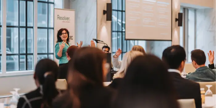 Keynote speaker engaging with raised hands from attendees at a grc day 2024 session, with a resolver banner in the background.