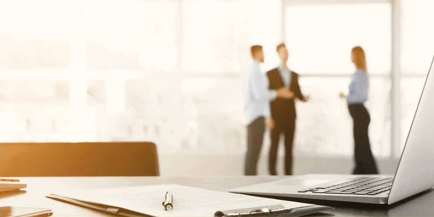 A workplace scene with a laptop and documents in the foreground and three blurred figures in discussion in the background, illustrating the importance of workplace violence prevention strategies.